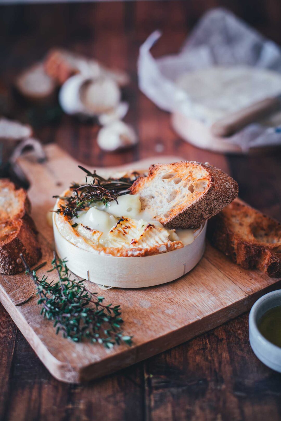 Nahaufnahme von einer Holzschachtel mit Camembert, der im Airfryer cremig zerlaufen ist. Getoppt mit Honig und Thymian und einem knusprigen Brot, das in den zerfließenden Käse getunkt wird. Im Hintergrund sieht man frischen Thymian, Knoblauch und weiteren Käse.