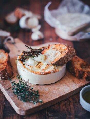 Nahaufnahme von einer Holzschachtel mit Camembert, der im Airfryer cremig zerlaufen ist. Getoppt mit Honig und Thymian und einem knusprigen Brot, das in den zerfließenden Käse getunkt wird. Im Hintergrund sieht man frischen Thymian, Knoblauch und weiteren Käse.