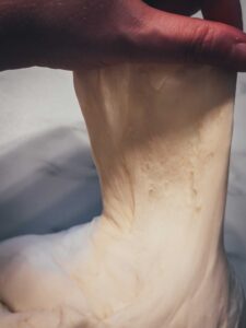 Nahaufnahme von Hefeteig, der mit der Hand für den Fenstertest hochgezogen wird – so sieht man, ob der Teig gut ausgeknetet ist.