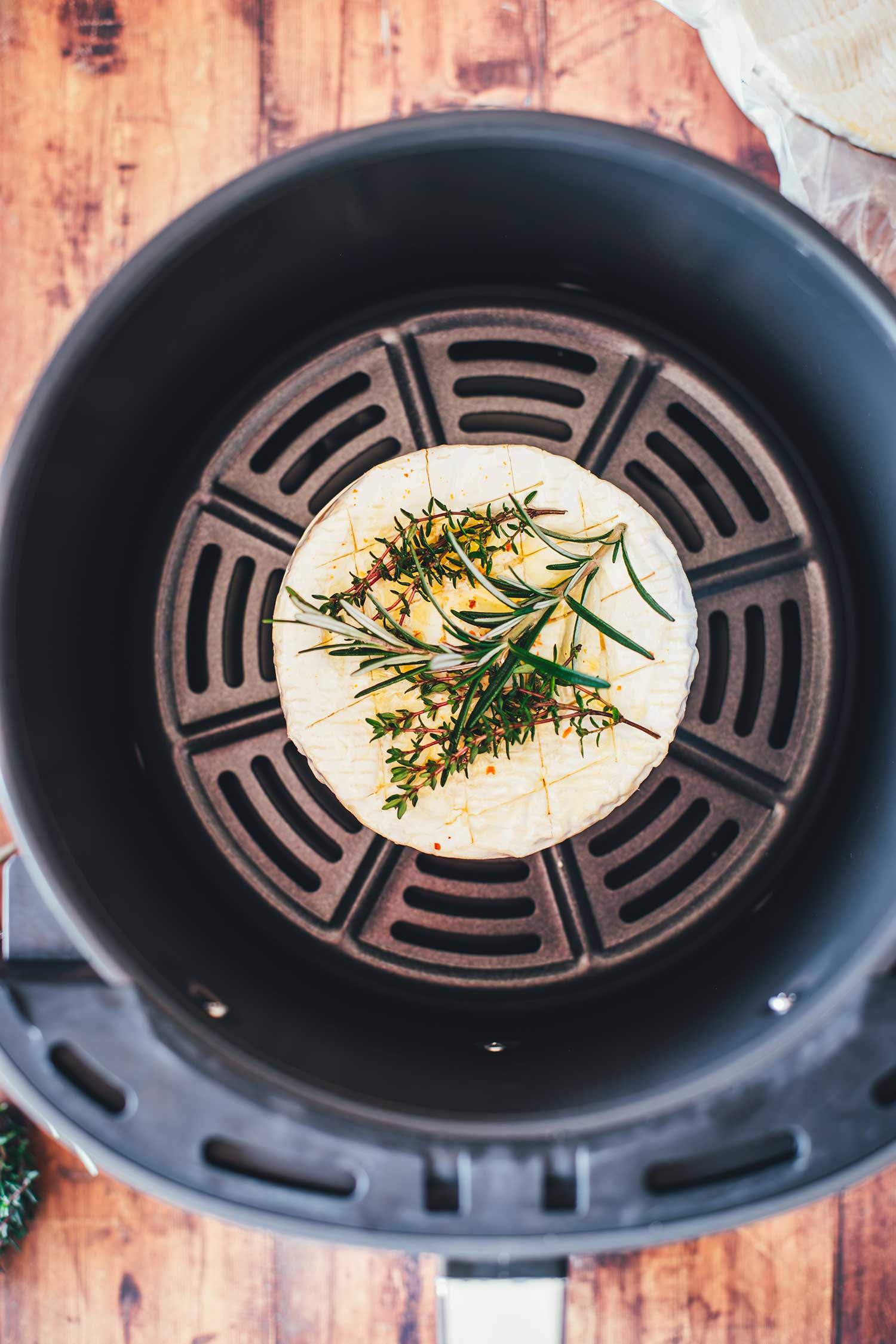 Vorbereiteter Camembert mit eingeritzter Rinde und frischen Kräutern im Korb der Heißluftfritteuse vor dem Backen.