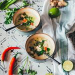 Zwei Schalen Thai-Kokos-Suppe mit Huhn, Pak Choi und Chili, serviert mit Limetten, Ingwer und Koriander auf einem rustikalen Untergrund | moeyskitchen.com