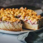 Frisch gebackener Rhabarber-Schmand-Kuchen liegt auf einer Kuchenplatte. Ein Stück wurde herausgeschnitten und liegt servierbereit auf einem Tortenheber. Man sieht den Kuchen im Ganzen und dazu die Schichten aus Rührteig, Schmandcreme, Rhabarber sowie die knusprigen Streusel.