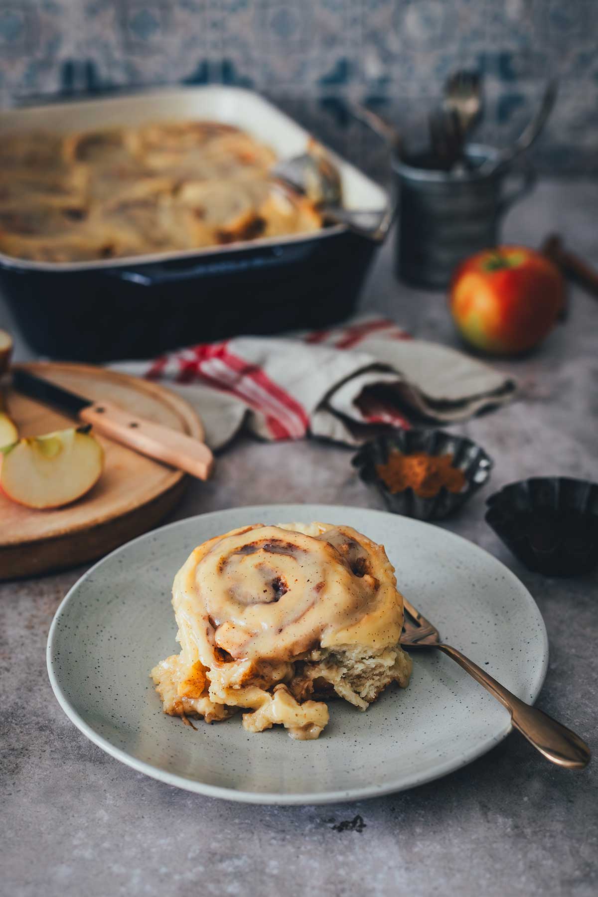 Seitliche Ansicht einer einzelnen Apfel-Zimtschnecke auf dem Teller. Im Hintergrund steht die Backform mit den restlichen Apfel-Zimtschnecken | moeyskitchen.com