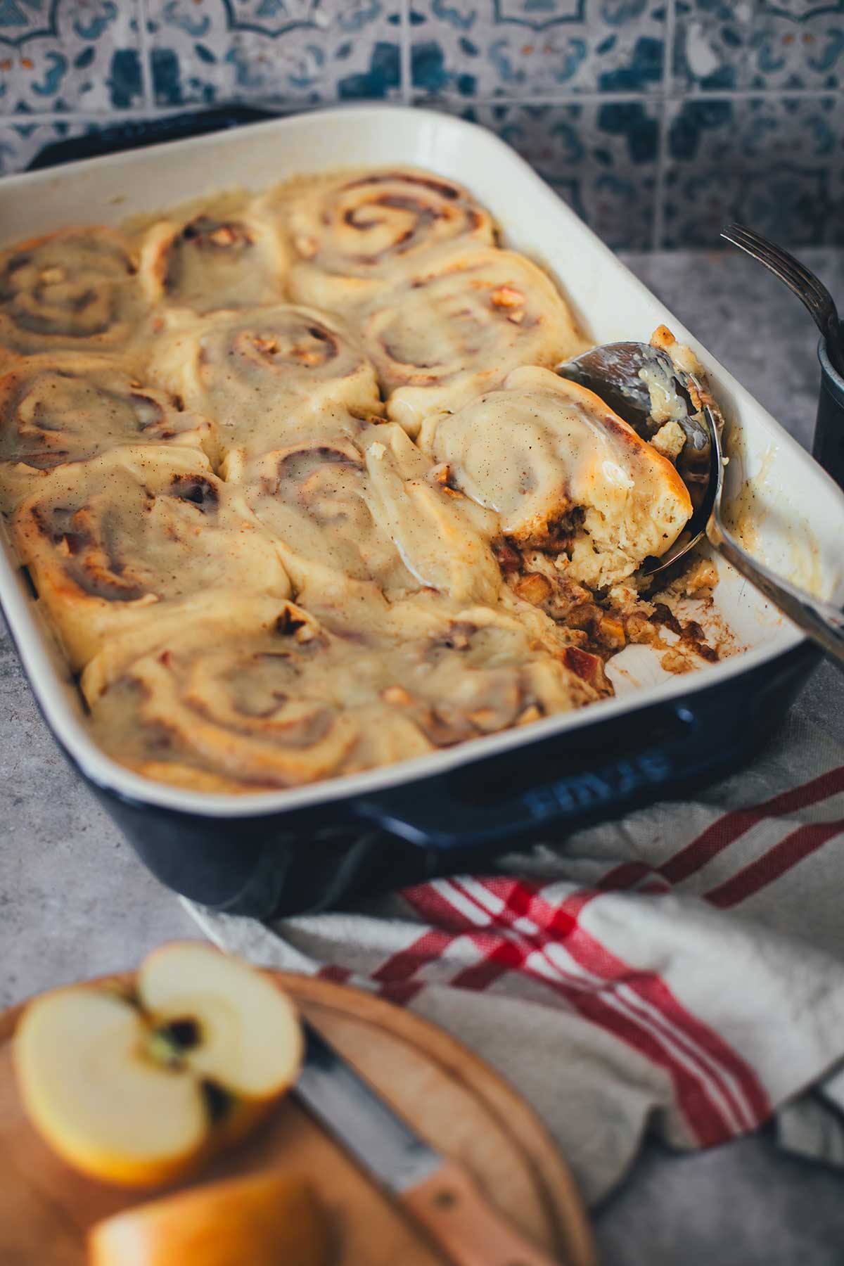 Seitliche Aufnahme einer Backform, gefüllt mit Apple Cinnamon Rolls. Eine Rolle fehlt, dafür liegt ein Löffel mit in der Form. Im Vordergrund liegt ein halber Apfel | moeyskitchen.com