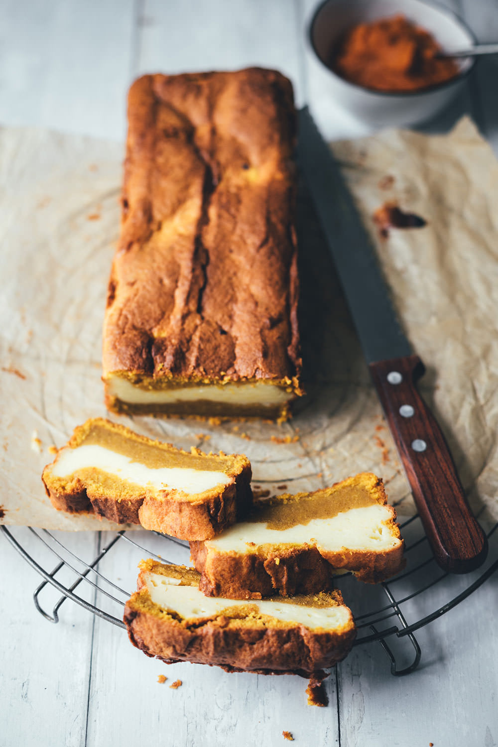 Kürbis-Frischkäse-Kuchen ist ein Pumpkin Bread mit Cheesecake – hier als Kastenkuchen auf einem Stück Backpapier mit einem Brotmesser. Drei Scheiben sind abgeschnitten und man erkennt die einzelnen Schichten. | moeyskitchen.com