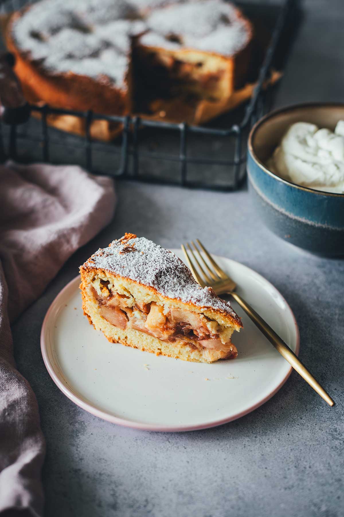 Ein Stück Appeltaart auf einem Teller, im Hintergrund sieht man den ganzen Apfelkuchen und daneben eine Schüssel mit Schlagsahne | moeyskitchen.com