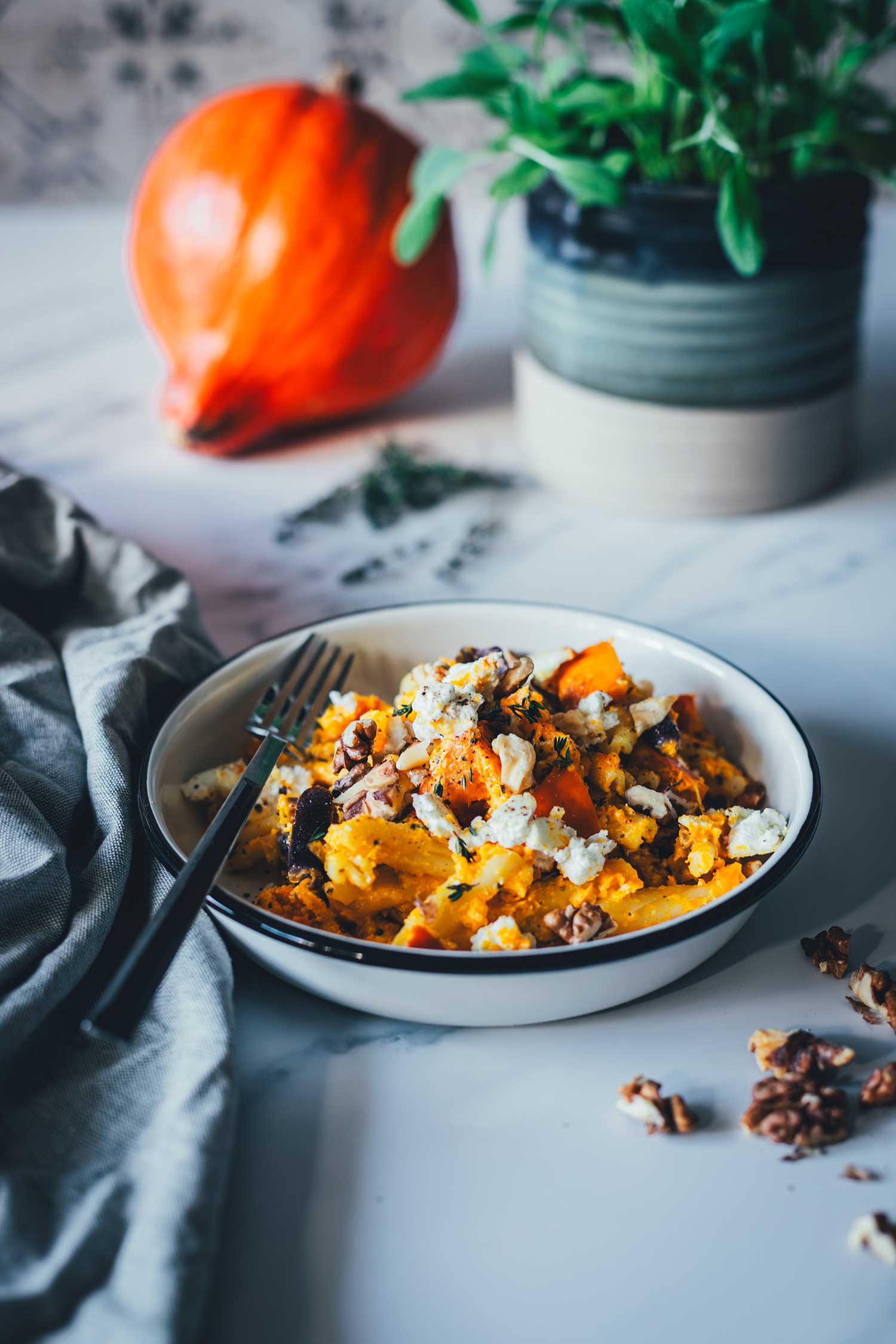 Seitliche Ansicht eines Tellers mit einem Pastagericht – cremige Kürbis-Feta-Pasta, bestreut mit Walnüssen. Im Hintergrund liegt ein Hokkaido-Kürbis, daneben ein Topf mit Salbei. Im Vordergrund sieht man eine Serviette und Walnusskerne. | moeyskitchen.com
