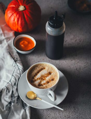 Eine Tasse Cappuccino, getoppt mit Pumpkin Spice Sirup mit brauner Butter, der in eine Squeezeflasche daneben steht. Im Hintergrund sieht man einen Hokkaido Kürbis und Kürbispüree in einer kleinen Schüssel. | moeyskitchen.com