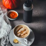 Eine Tasse Cappuccino, getoppt mit Pumpkin Spice Sirup mit brauner Butter, der in eine Squeezeflasche daneben steht. Im Hintergrund sieht man einen Hokkaido Kürbis und Kürbispüree in einer kleinen Schüssel. | moeyskitchen.com