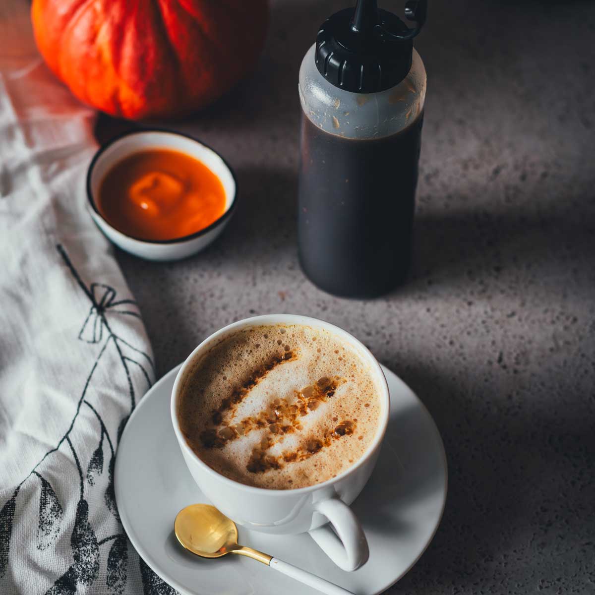 Eine Tasse Cappuccino, getoppt mit Pumpkin Spice Sirup mit brauner Butter, der in eine Squeezeflasche daneben steht. Im Hintergrund sieht man einen Hokkaido Kürbis und Kürbispüree in einer kleinen Schüssel. | moeyskitchen.com