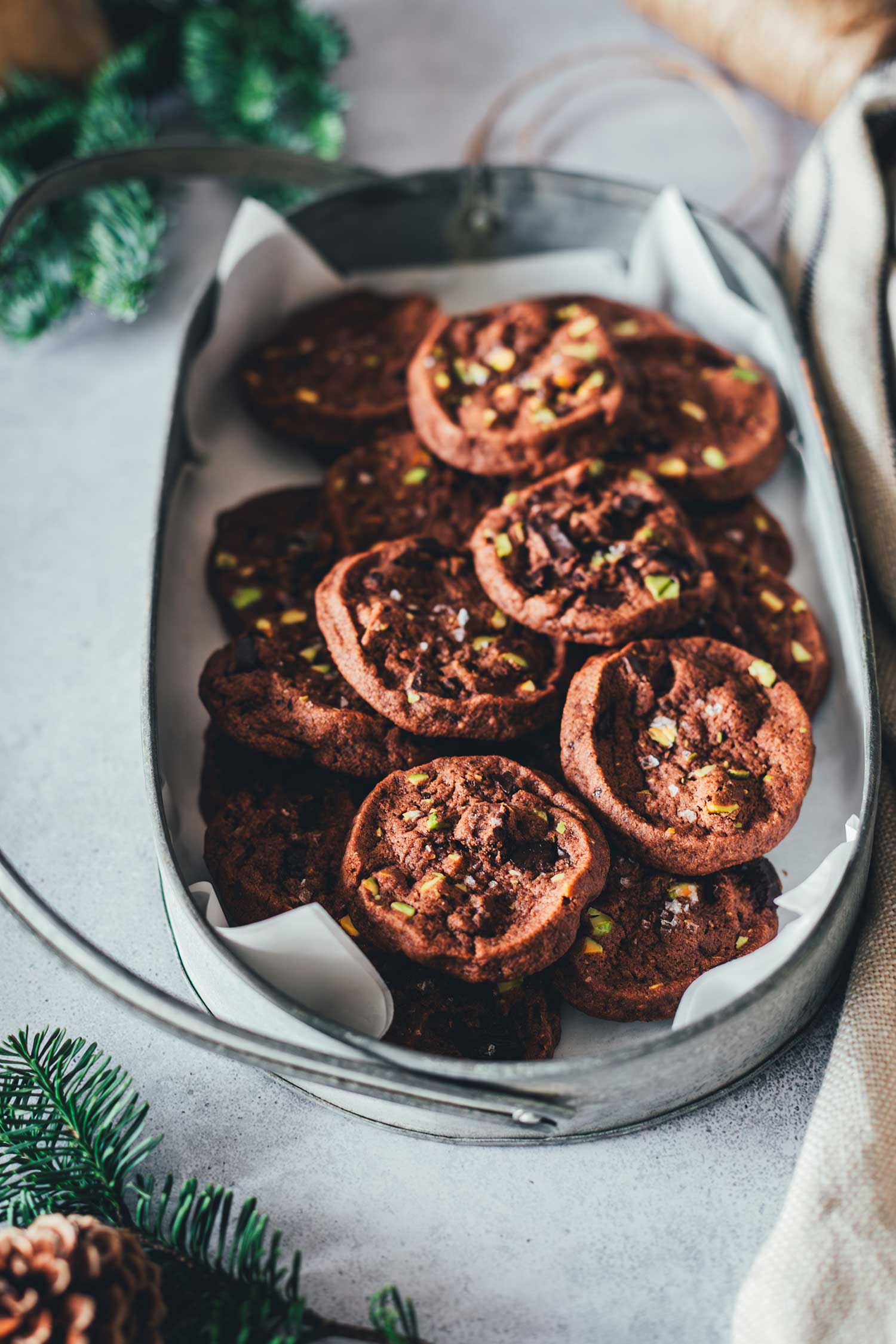 Seitliche Ansicht von einem Tablett mit braunen Schokoladen-Pistazien-Cookies gefüllt, drumherum mit Tannengrün und Geschirrtuch dekoriert
