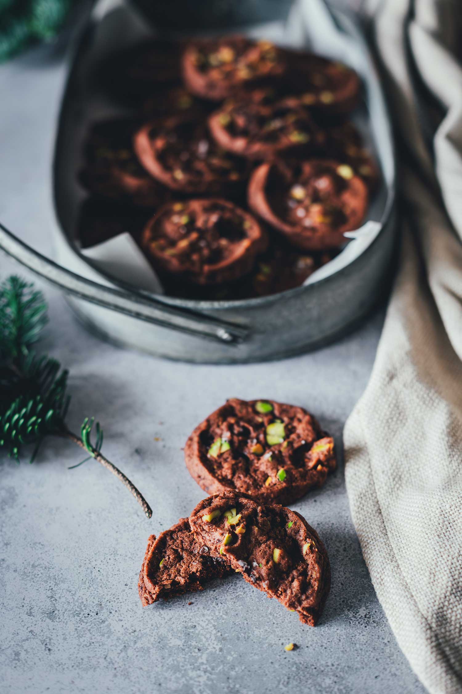 Nahaufnahme von zwei Schokoladen-Pistazien-Cookies, einer ist in der Mitte durchgebrochen. Im Hintergrund sieht man etwas Tannengrün und weitere Cookies