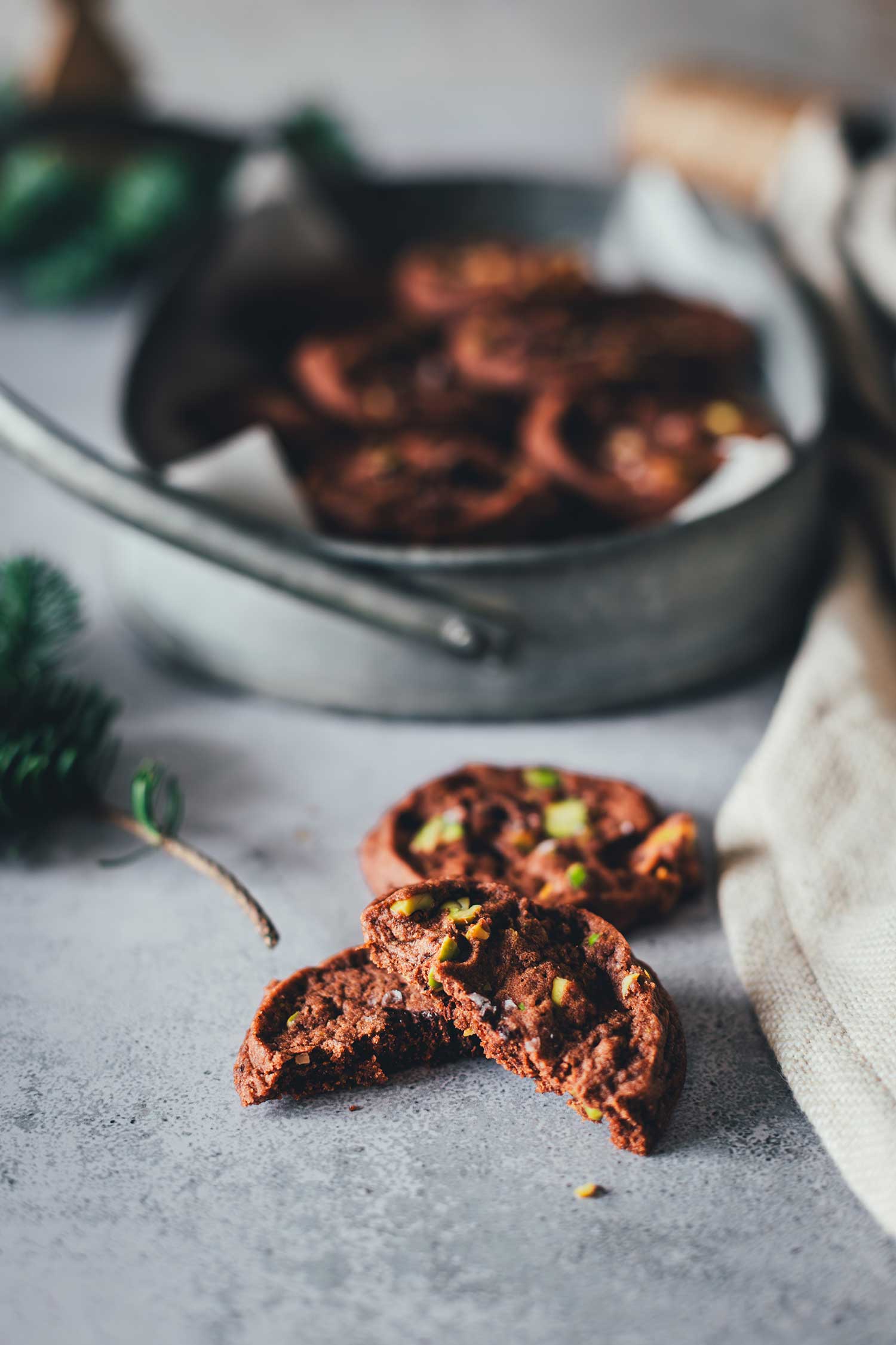 Nahaufnahme von zwei Schokoladen-Pistazien-Cookies, einer ist in der Mitte durchgebrochen. Im Hintergrund sieht man etwas Tannengrün und weitere Cookies