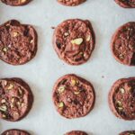 Topshot Bild von braunen Schokoladen-Pistazien-Cookies mit Meersalz bestreut auf einem Backblech mit weißem Backpapier liegend