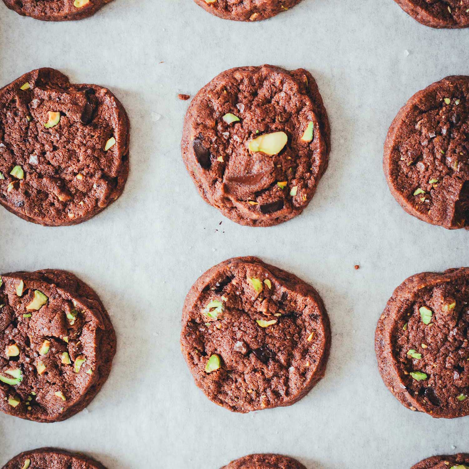 Topshot Bild von braunen Schokoladen-Pistazien-Cookies mit Meersalz bestreut auf einem Backblech mit weißem Backpapier liegend