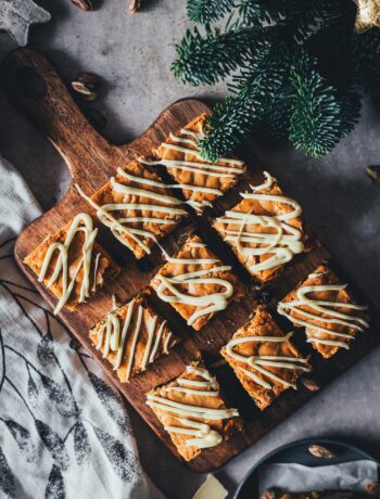 Weihnachts-Blondies mit weißer Schokolade, Cranberries und Pistazien auf einem Holzbrett, von oben fotografiert.