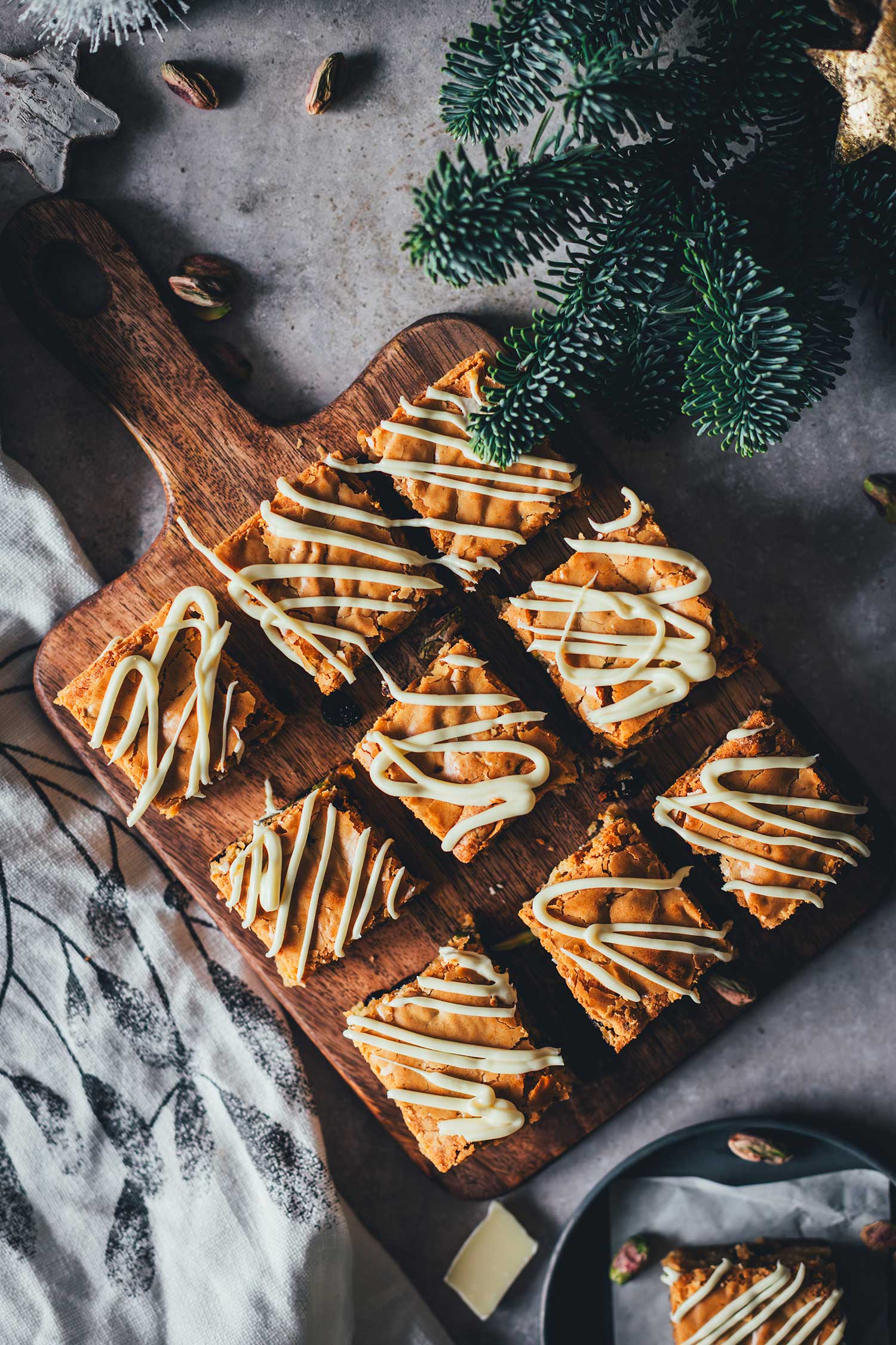 Weihnachts-Blondies mit weißer Schokolade, Cranberries und Pistazien auf einem Holzbrett, von oben fotografiert.