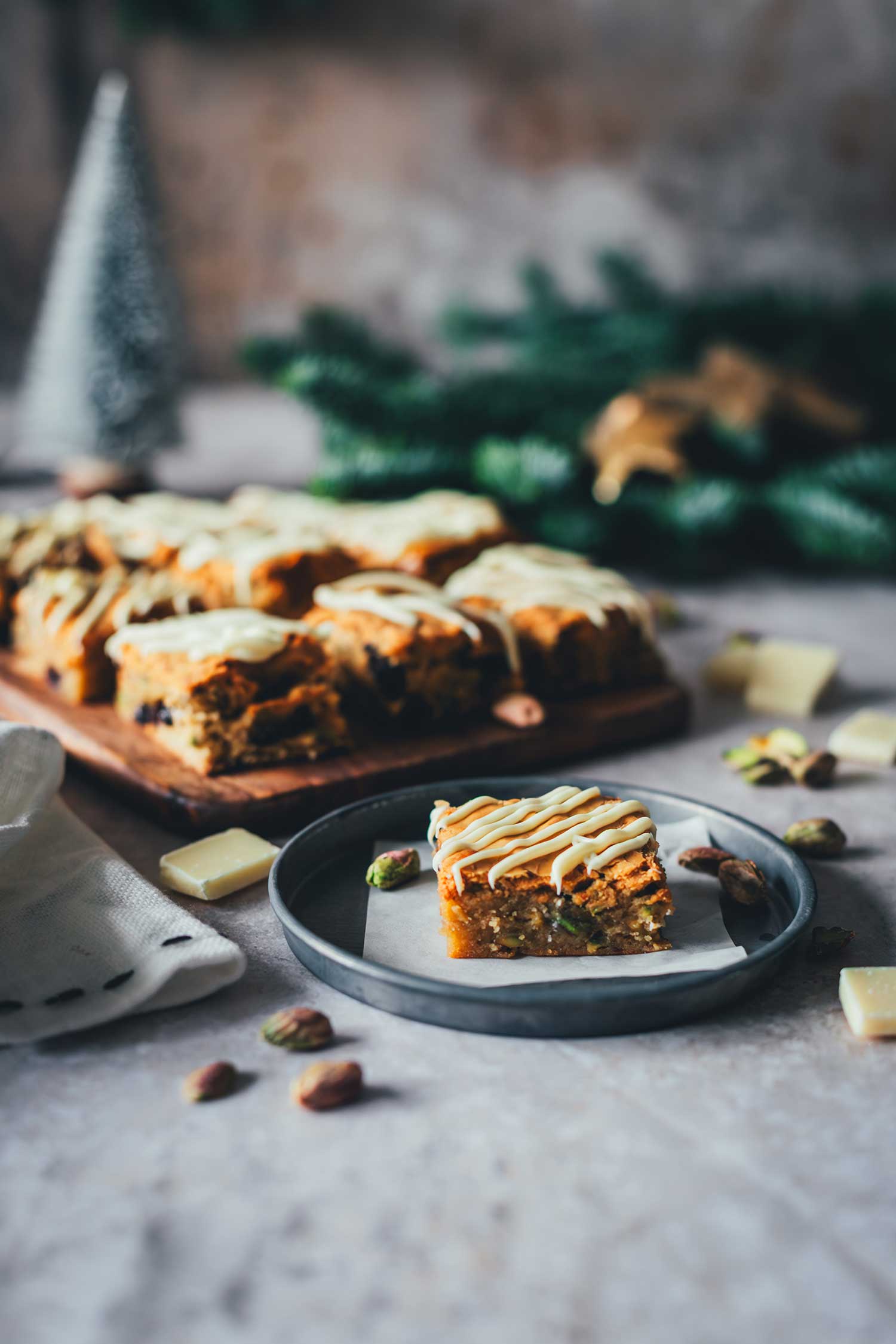 Ein Stück Weihnachts-Blondie auf einem Teller mit weiteren Blondies im Hintergrund im weihnachtlichen Setting.