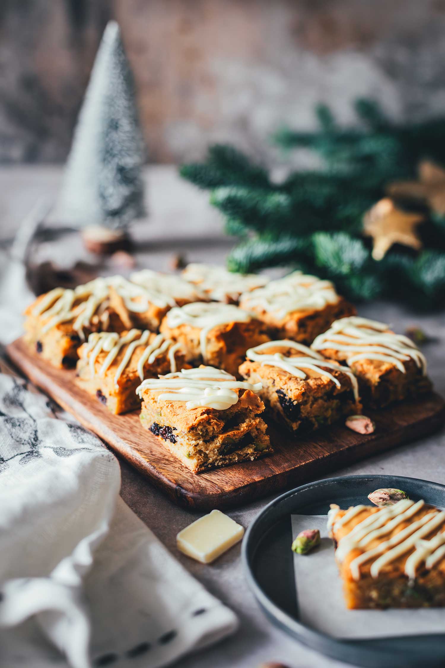 Ein Stück Weihnachts-Blondie auf einem Teller mit weiteren Blondies im Hintergrund im weihnachtlichen Setting.