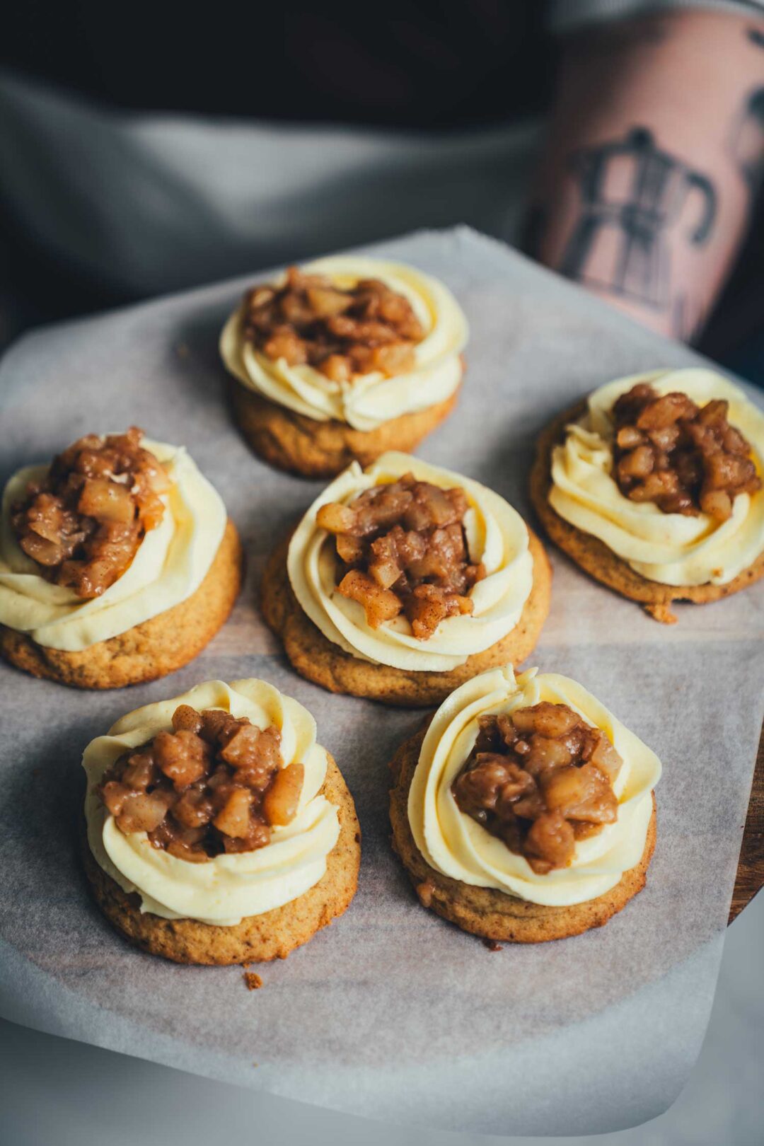 Brown Butter Apple Pie Cookies auf einem Tablett, frisch dekoriert mit Mascarpone-Frosting und Apfeltopping; im Hintergrund unscharfer Arm einer Person beim Servieren.