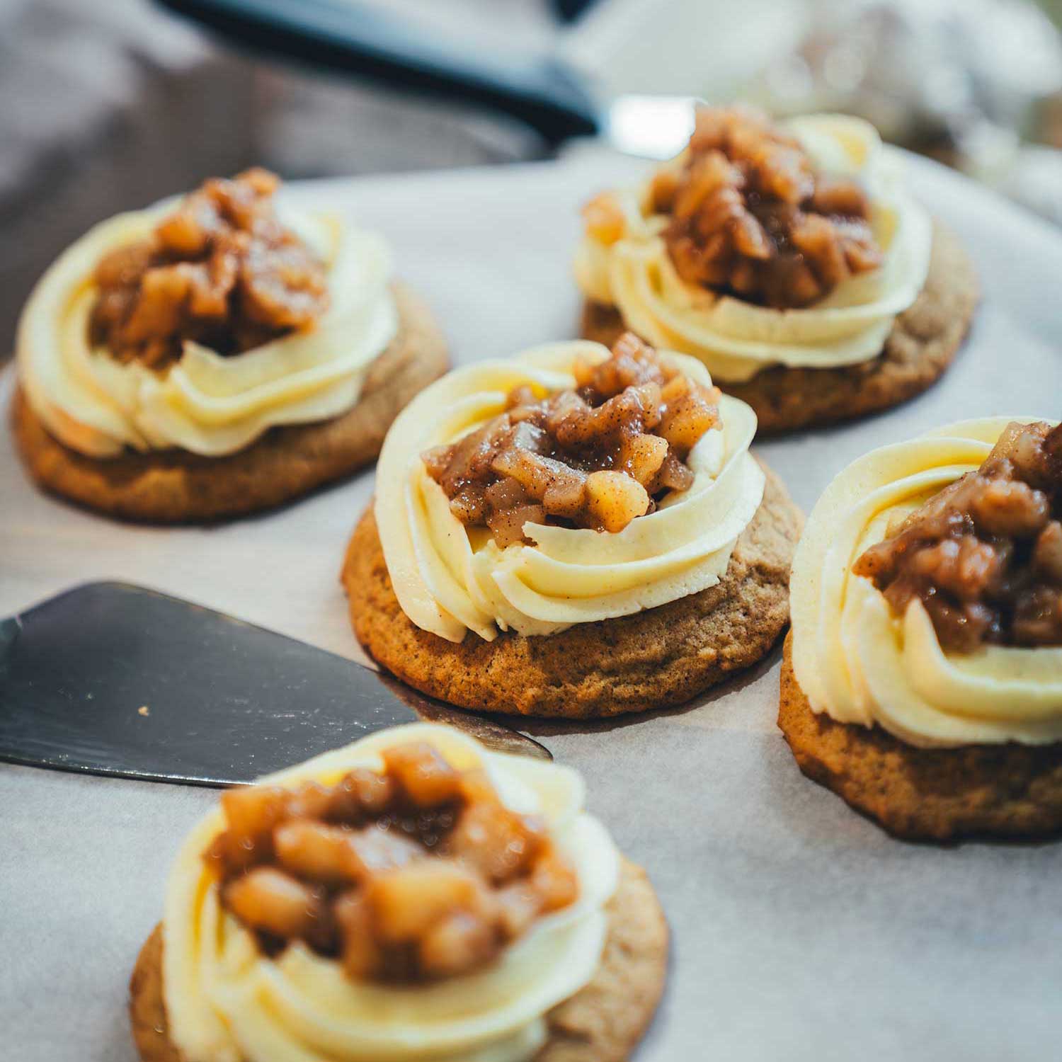 seitliche Ansicht von Apple Pie Cookies mit Mascarpone-Frosting und Apfeltopping auf einer Tortenplatte mit weißem Backpapier