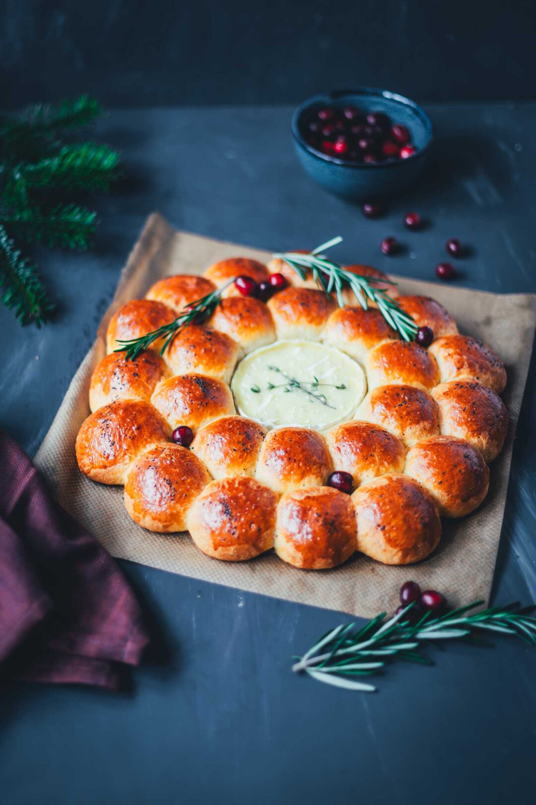 Herzhafter Brioche-Kranz mit geschmolzenem Camembert in der Mitte, dekoriert mit Rosmarin und Cranberries auf dunklem Hintergrund – festlicher Snack für die Adventszeit.