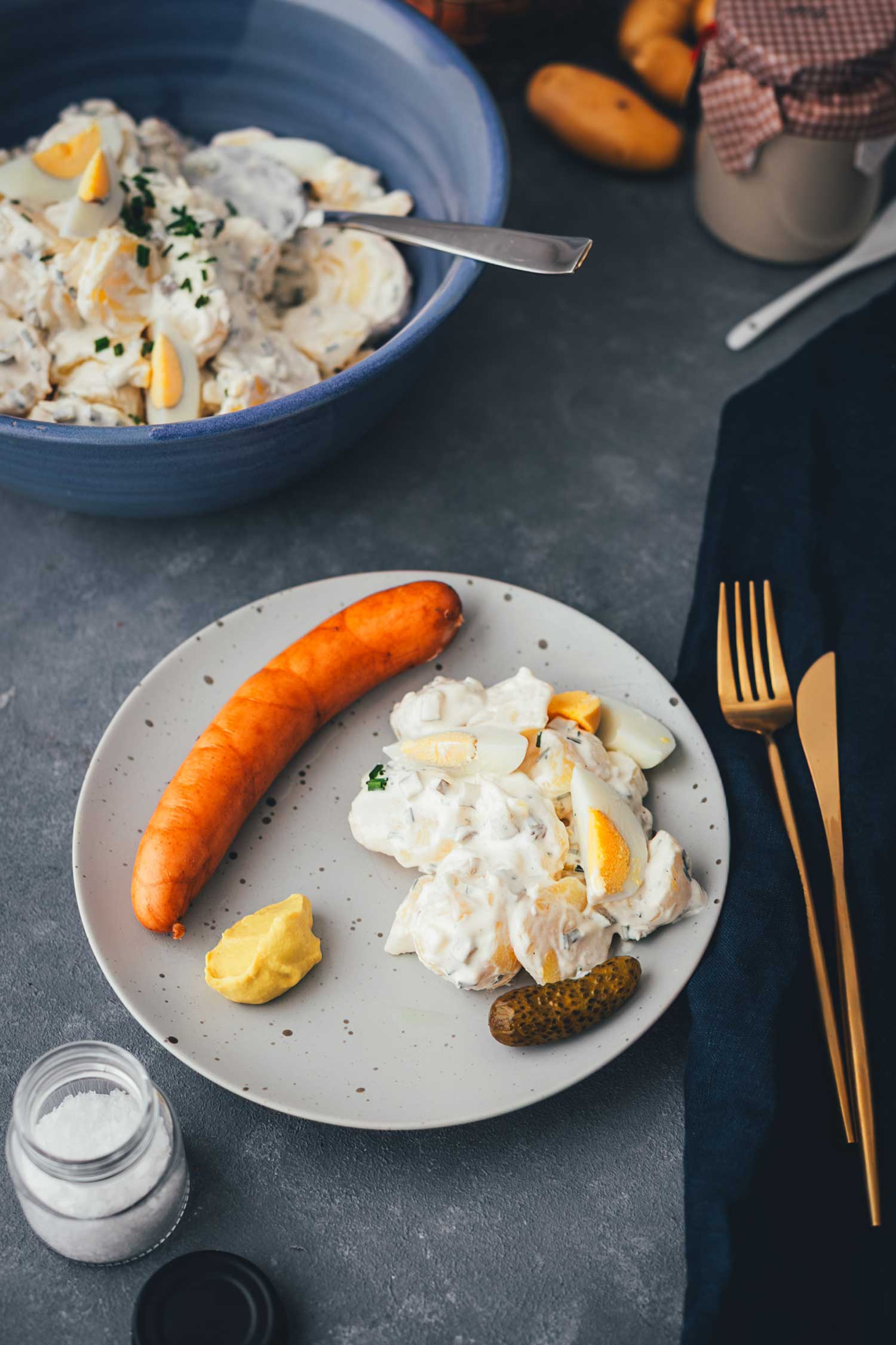 Teller mit cremigem Kartoffelsalat, Bockwurst, Senf und Cornichon, daneben eine große Schüssel Kartoffelsalat – klassisches Weihnachtsessen.