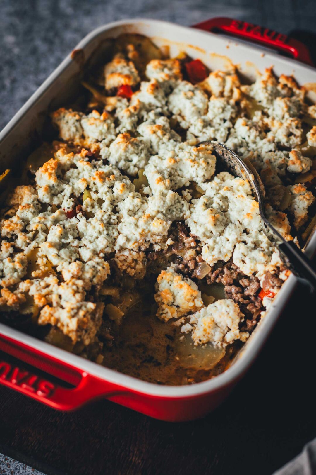 Frisch gebackener Kartoffel-Hack-Auflauf mit einer knusprigen Feta-Kruste in einer roten Keramik-Auflaufform, aus der bereits eine Portion mit einem Löffel entnommen wurde.