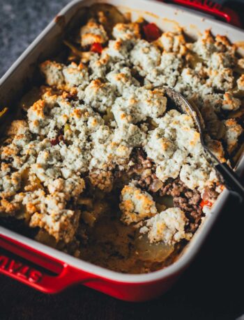 Frisch gebackener Kartoffel-Hack-Auflauf mit einer knusprigen Feta-Kruste in einer roten Keramik-Auflaufform, aus der bereits eine Portion mit einem Löffel entnommen wurde.