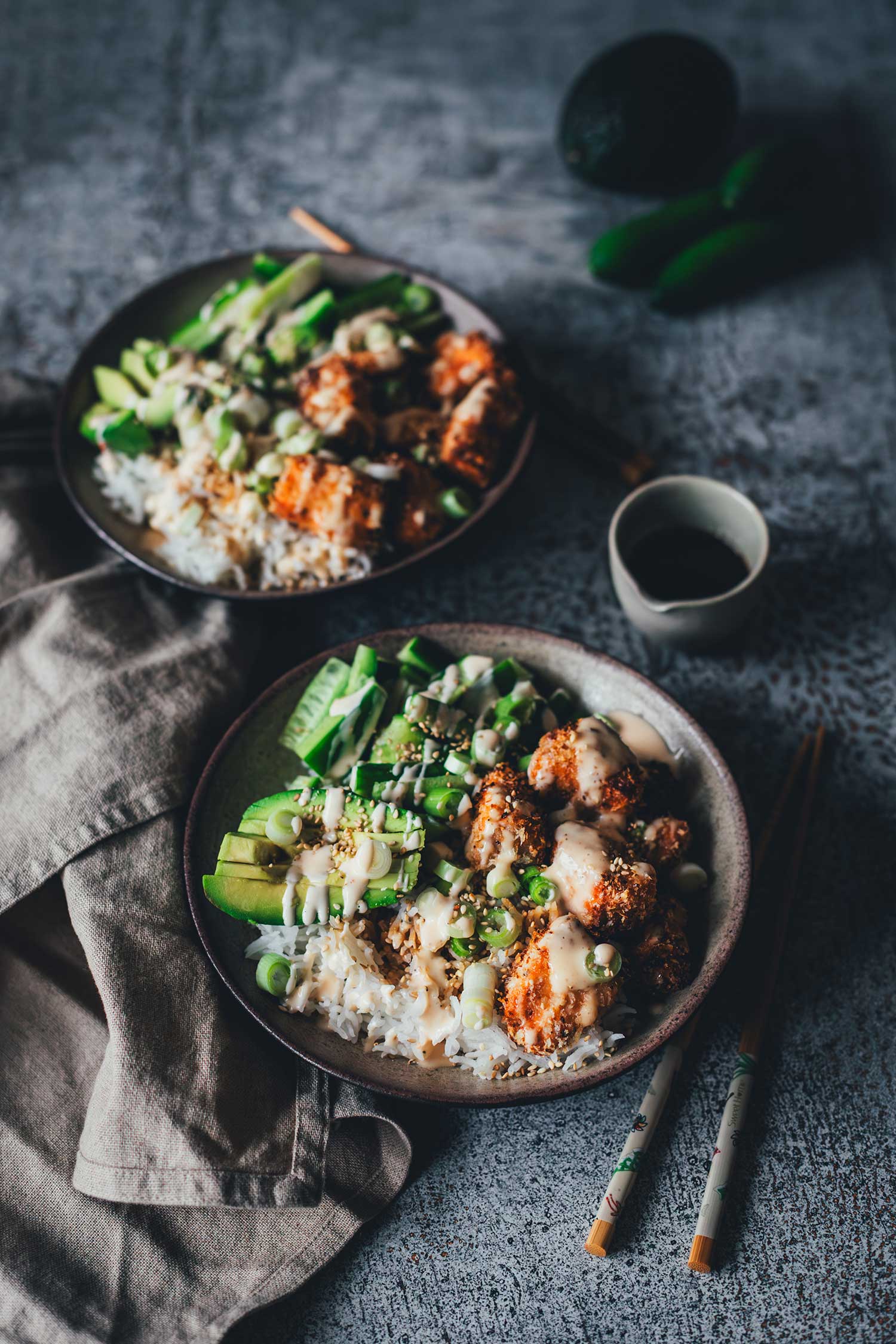 Schräge Ansicht von oben auf zwei fertig angerichtete Crispy Lachs Bowls mit Avocado, Gurke und Stäbchen in einer gemütlichen Food-Setting-Atmosphäre