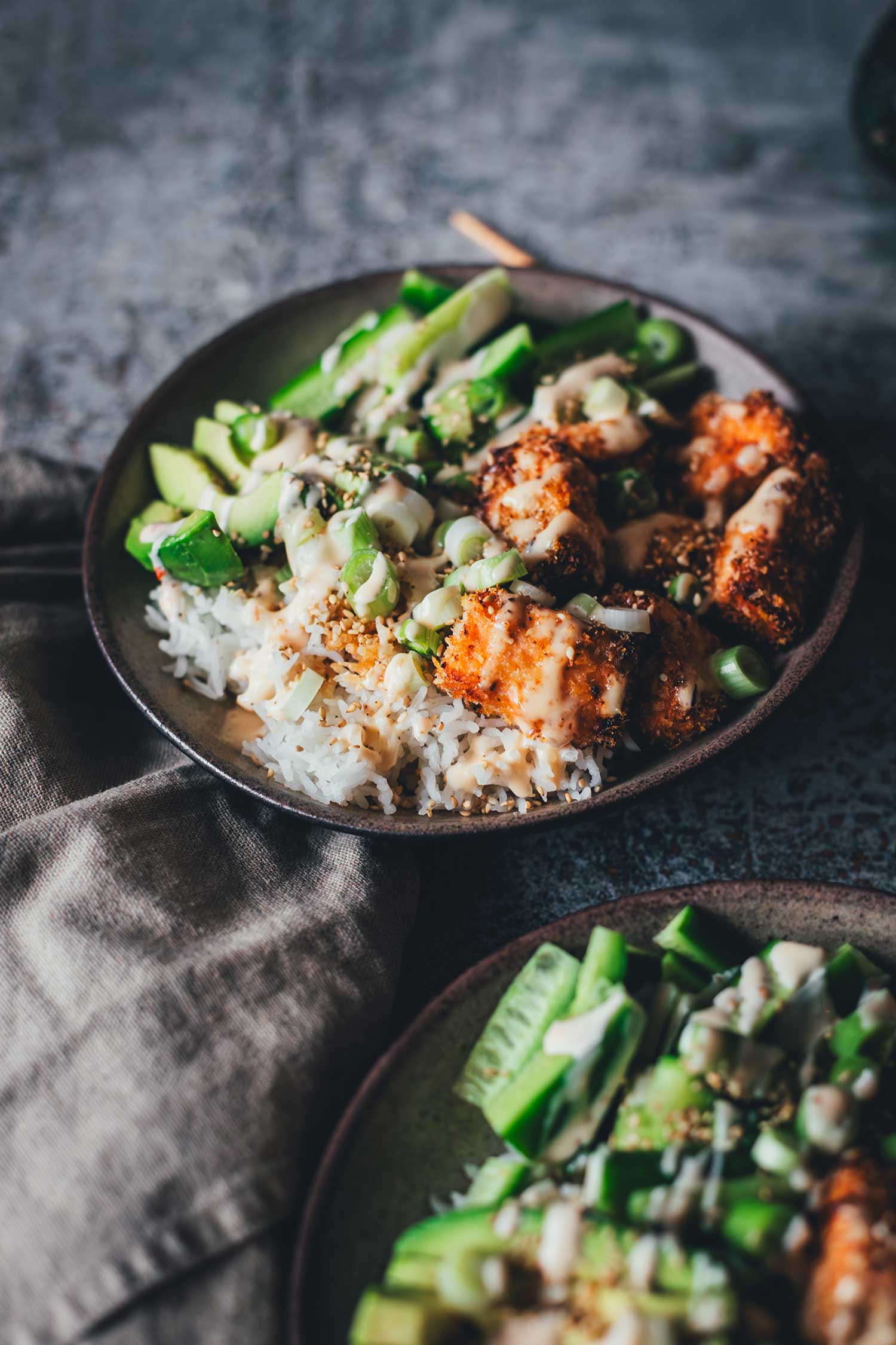 Nahaufnahme einer Crispy Lachs Bowl mit Fokus auf der oberen Schüssel, angerichtet mit Reis, Avocado, Frühlingszwiebeln und knusprigem Lachs