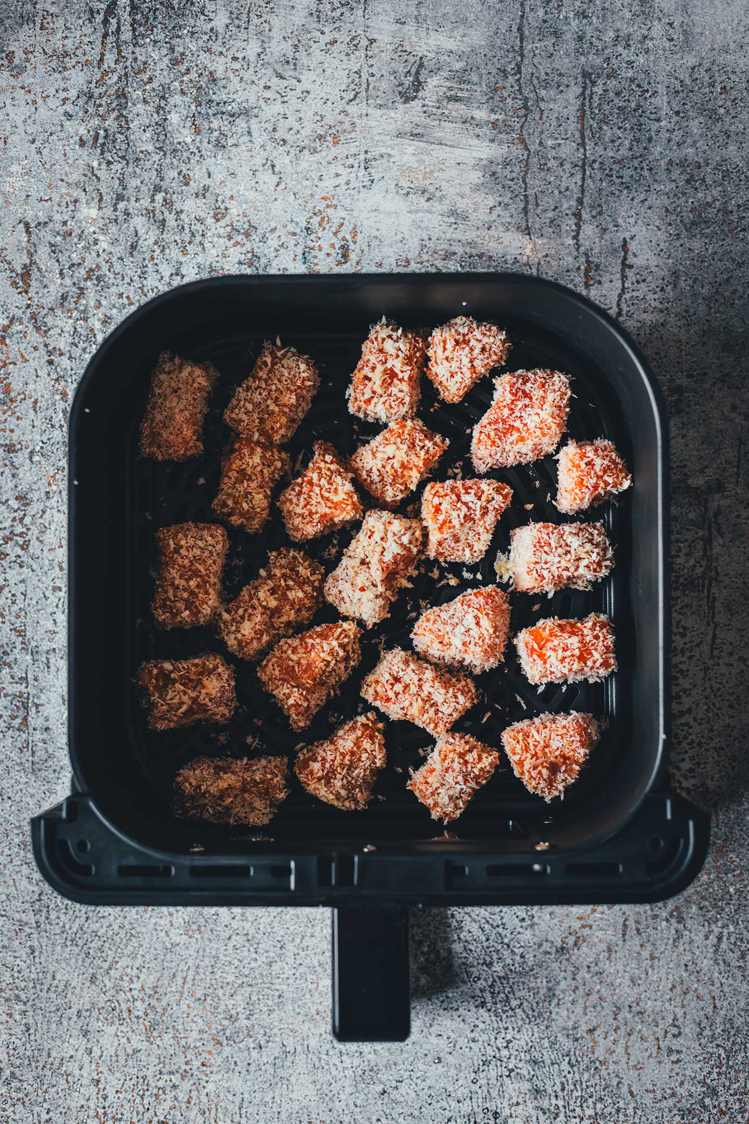 In Panko panierte Lachswürfel nebeneinander im Frittierkorb der Heißluftfritteuse vor dem Backen