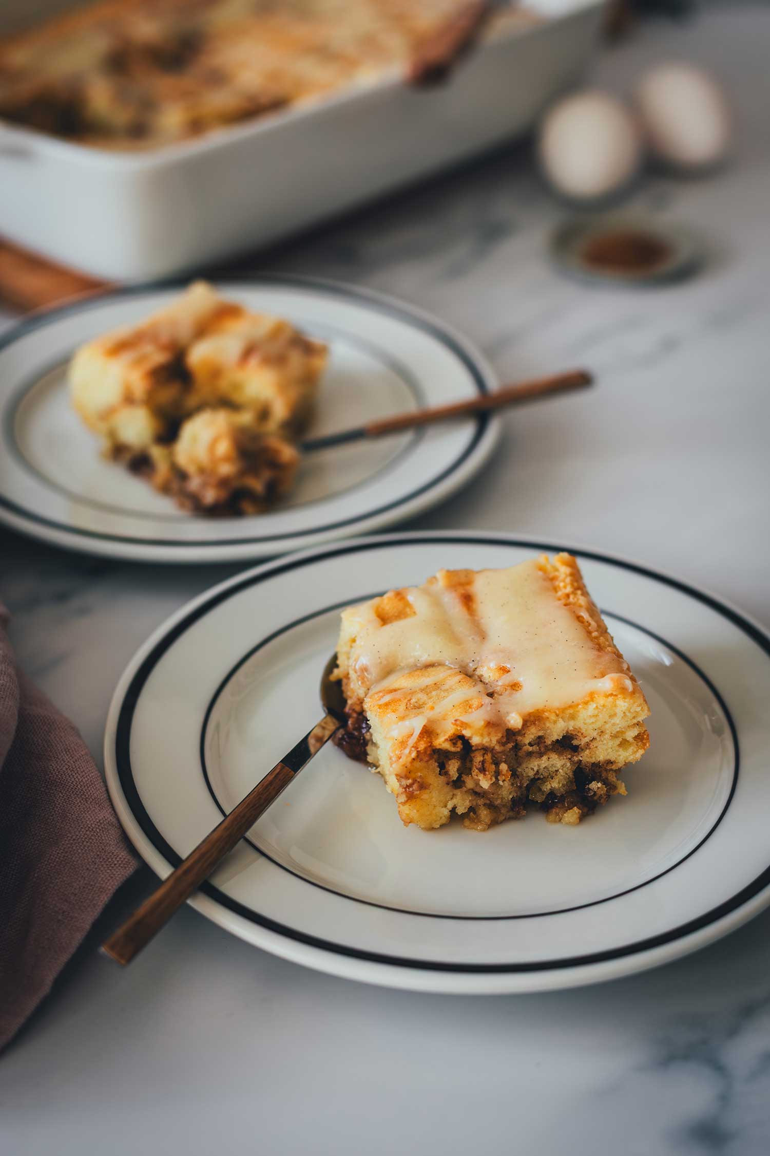 Zwei Kuchenstücke vom Zimtschnecken-Kuchen serviert auf zwei Tellern, im Hintergrund sieht man die Auflaufform.