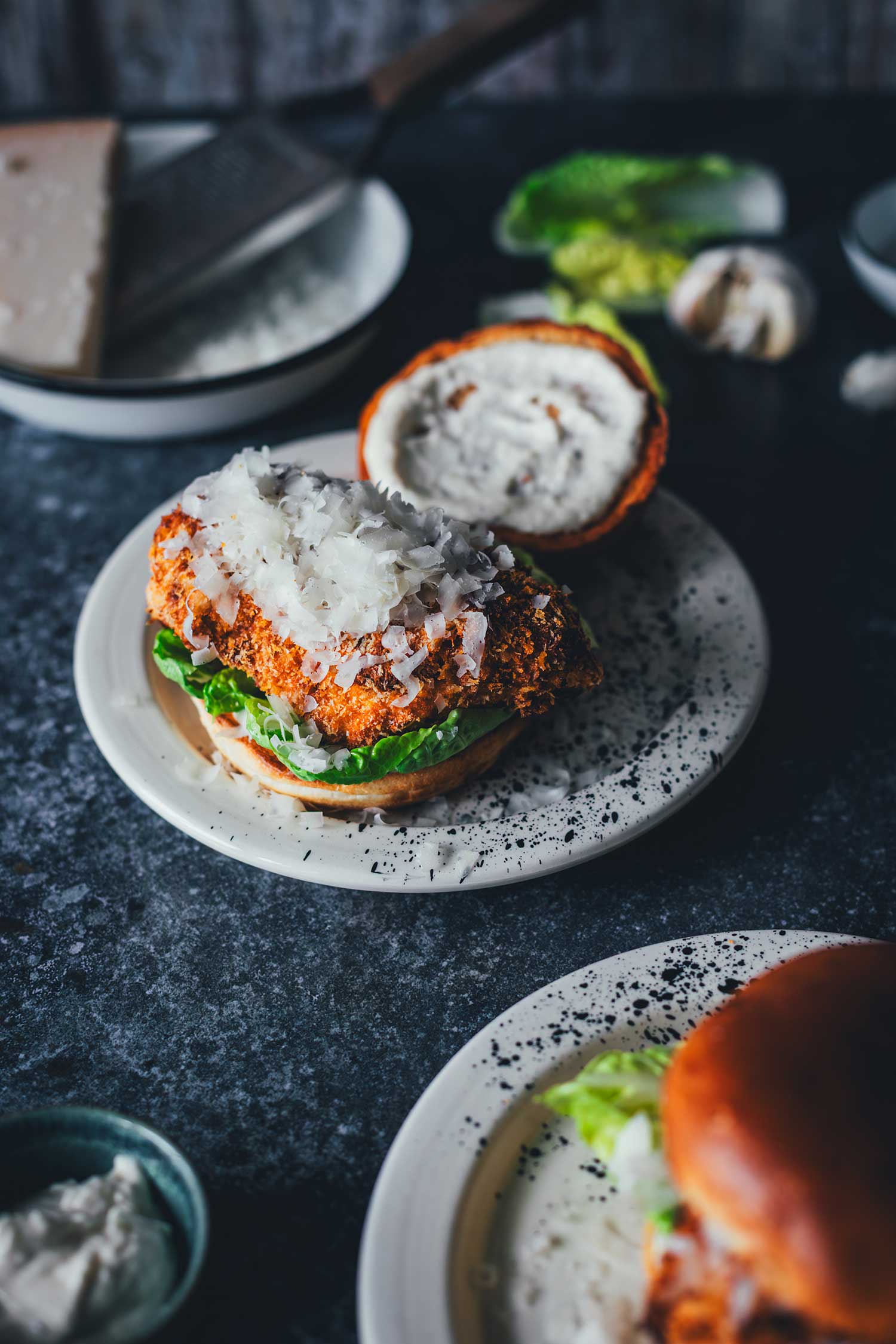 Hähnchen Burger ohne Deckel mit Panko-Kruste, Romanasalat und reichlich Parmesan auf getoastetem Brioche-Bun, im Hintergrund Microplane-Reibe und Käse