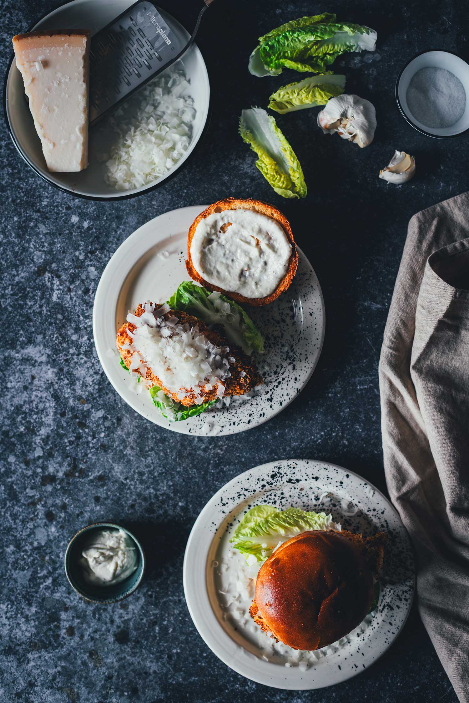 Crispy Chicken Burger Caesar-Style von oben fotografiert, dazu Zutaten wie Romanasalat, Knoblauch, Parmesan-Stück mit Microplane und Salz