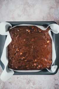 Brownie-Teig mit Macadamias und weißer Schokolade glatt gestrichen in einer mit Backpapier ausgelegten quadratischen Backform, bereit zum Backen.
