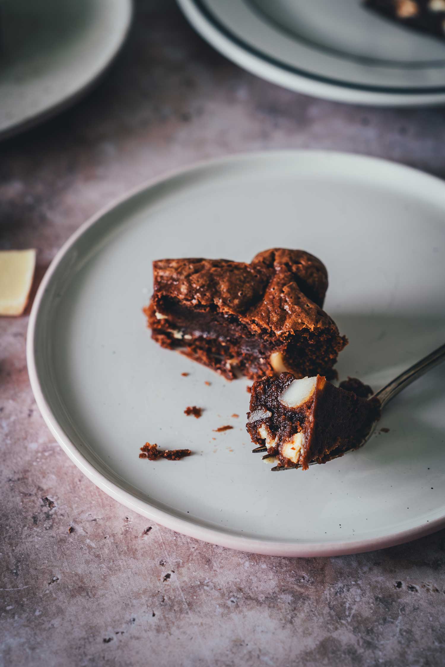 Angeschnittener fudgy Macadamia Brownie mit weißer Schokolade auf einem hellen Teller, Bissrest auf der Gabel.