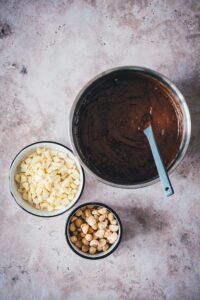 Geschmolzene Schokoladen-Butter-Mischung mit eingerührtem Zucker in einer Edelstahlschüssel, daneben weiße Schokolade und Macadamias.