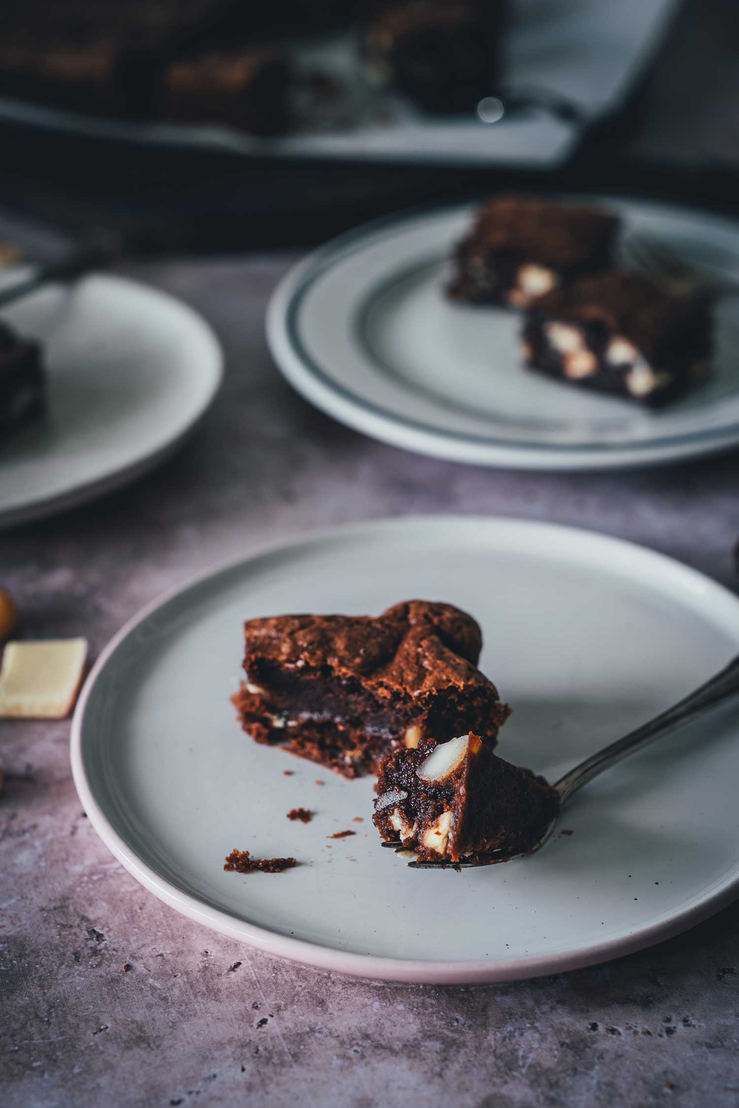 Saftiger Macadamia Brownie mit weißer Schokolade auf einem Teller, ein Stück mit der Gabel angehoben, mit sichtbarem Crinkle-Top.