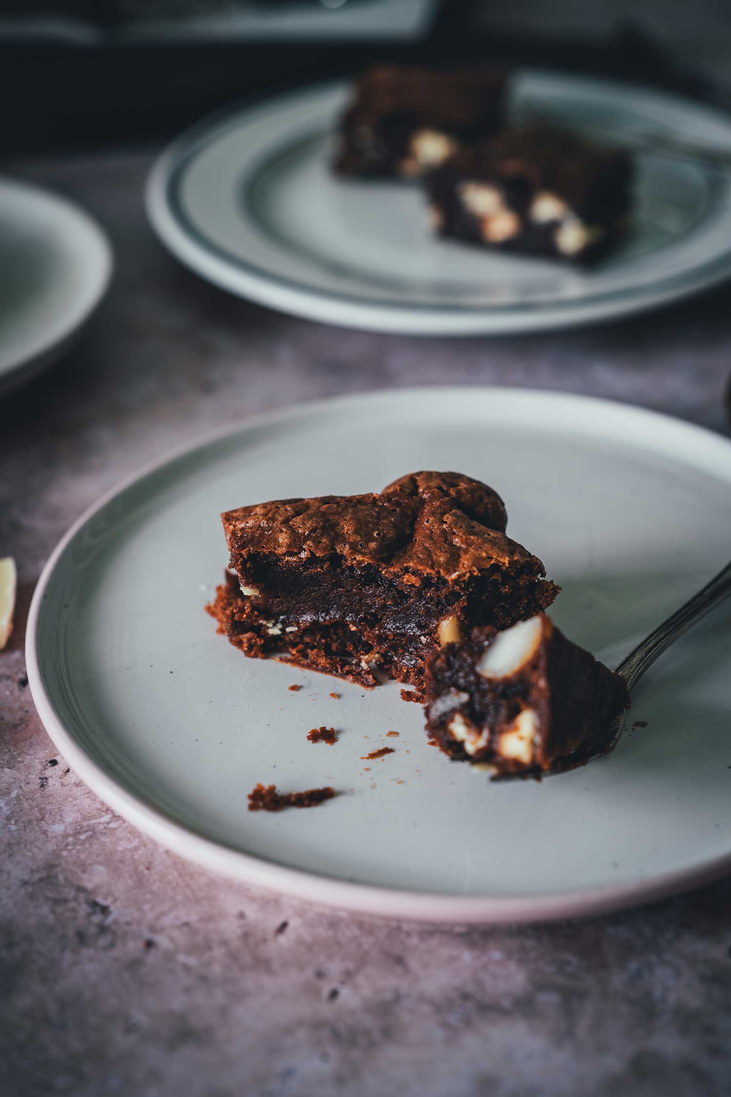 Macadamia Brownie mit weißer Schokolade auf einem weißen Teller, Bissrest auf der Gabel.