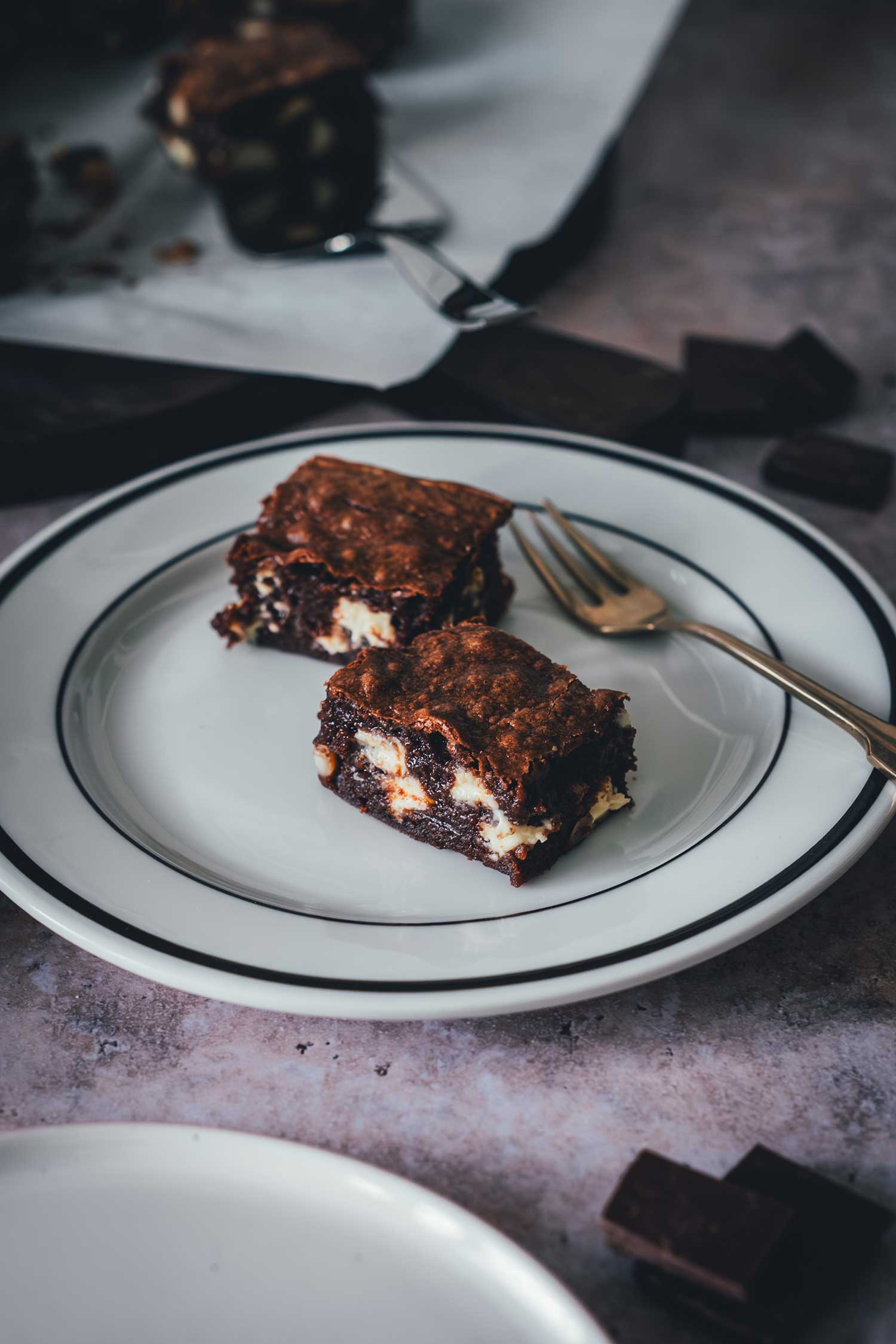 Zwei Macadamia Brownies mit weißer Schokolade auf einem Teller mit schwarzem Rand, Schokoladenstücke im Hintergrund.