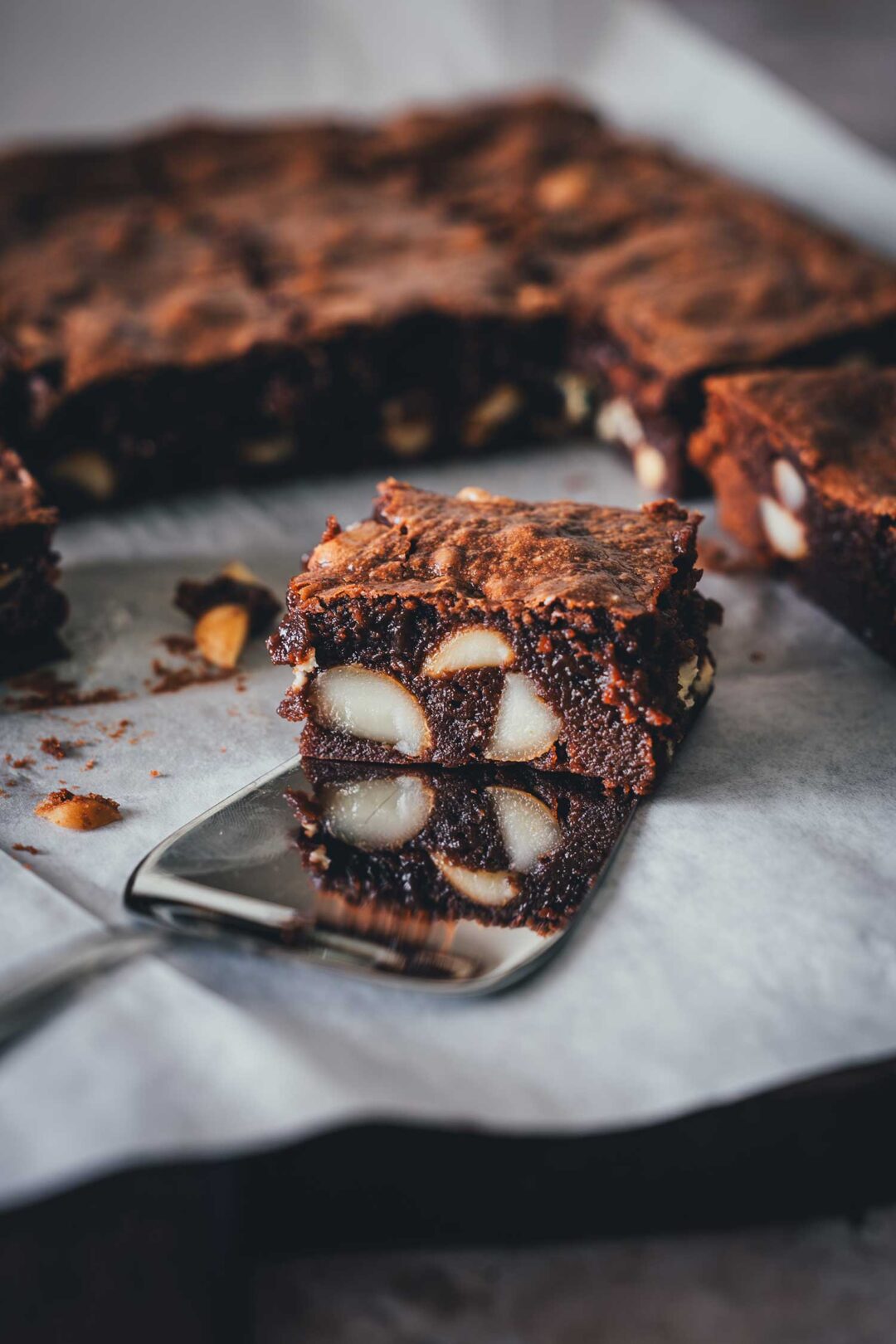 Saftiger Macadamia Brownie mit weißer Schokolade auf einem Tortenheber, mit sichtbarem Crinkle-Top und gestapeltem Anschnitt.
