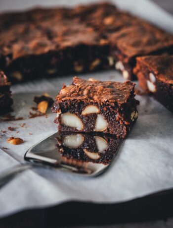Saftiger Macadamia Brownie mit weißer Schokolade auf einem Tortenheber, mit sichtbarem Crinkle-Top und gestapeltem Anschnitt.