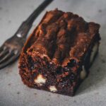 Saftiger Macadamia Brownie mit weißer Schokolade und Crinkle-Top auf einem grauen Teller mit Gabel.