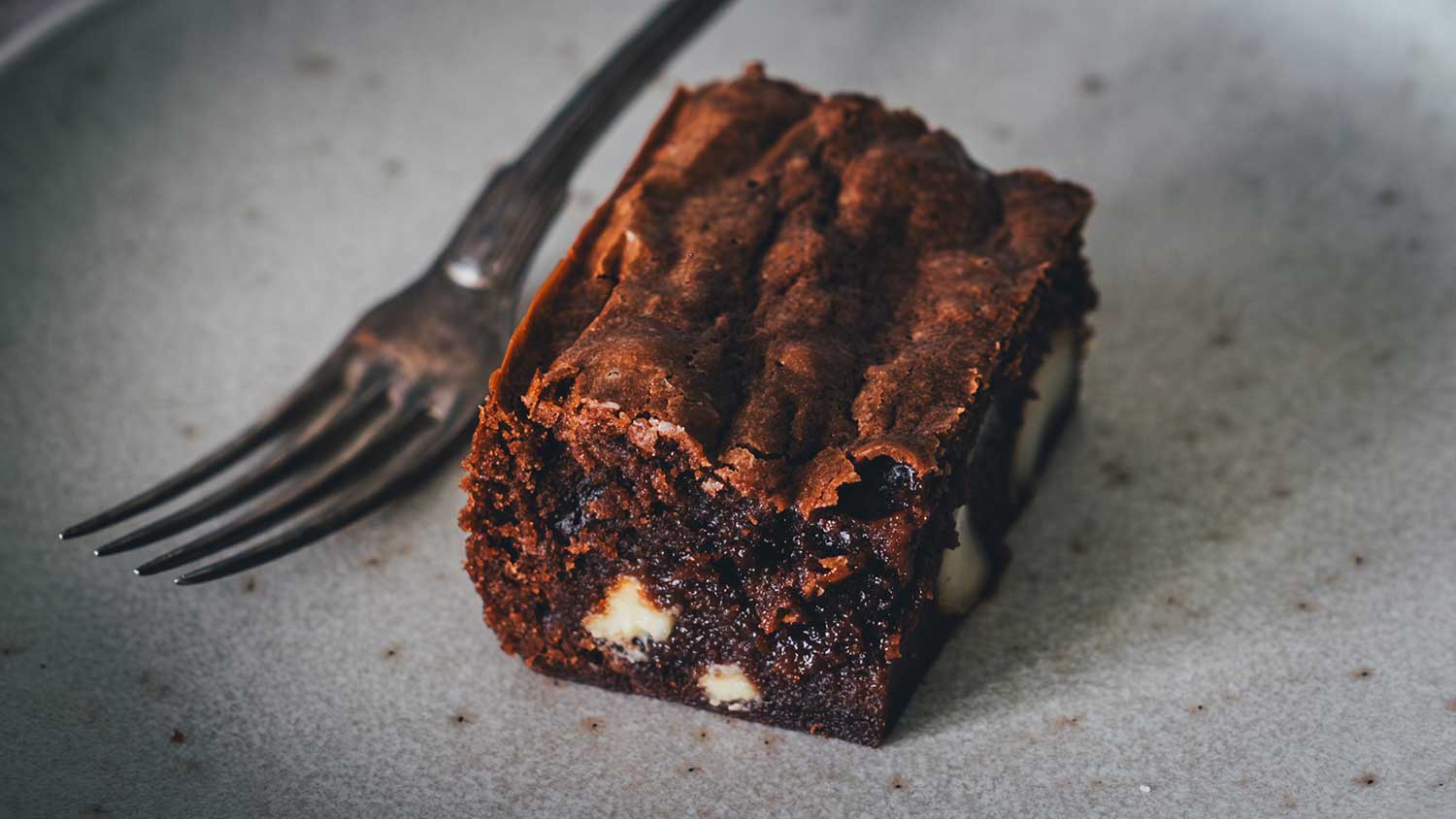 Saftiger Macadamia Brownie mit weißer Schokolade und Crinkle-Top auf einem grauen Teller mit Gabel.