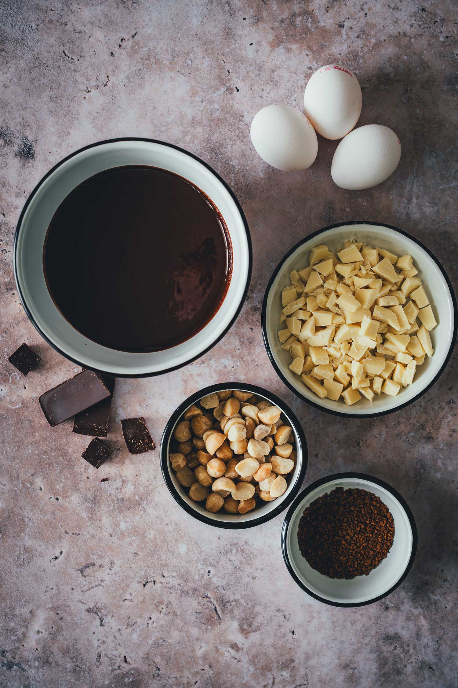 Zutaten für Macadamia Brownies mit weißer Schokolade: geschmolzene Edelbitterschokolade, weiße Schokolade, geröstete Macadamias, Espressopulver und Eier von oben.