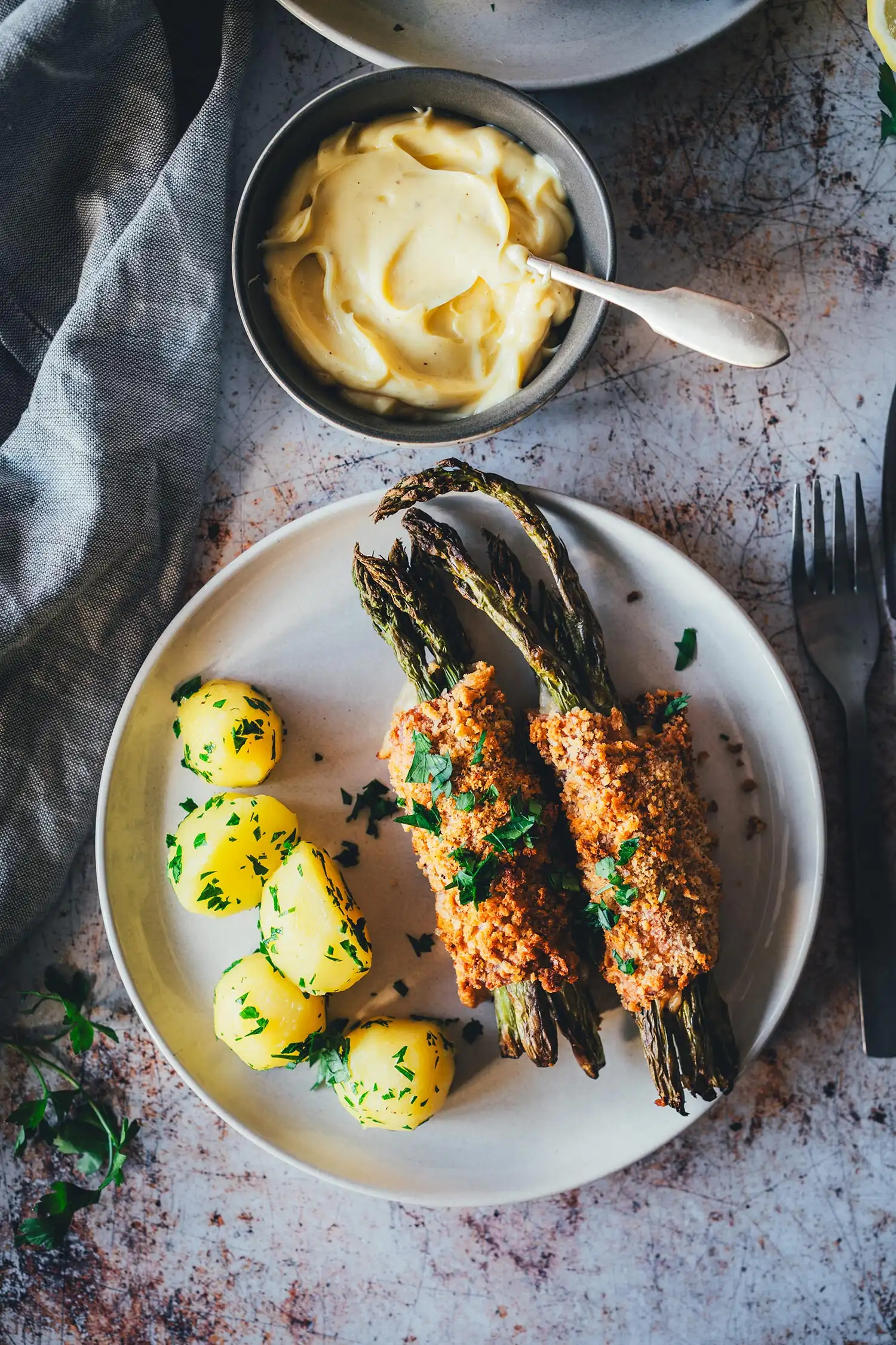 Angerichtetes Spargel Cordon Bleu auf hellem Teller mit Petersilienkartoffeln und Hollandaise im Hintergrund