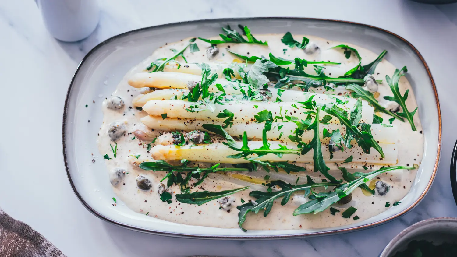 Spargel Tonnato mit weißem Spargel auf cremiger italienischer Thunfischsauce, garniert mit Rucola, Kapern und Petersilie auf ovaler Servierplatte