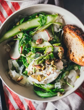 Grüner Spargelsalat mit Burrata, Pistazien und Rucola in einer hellen Schale, daneben getoastetes Sauerteigbrot