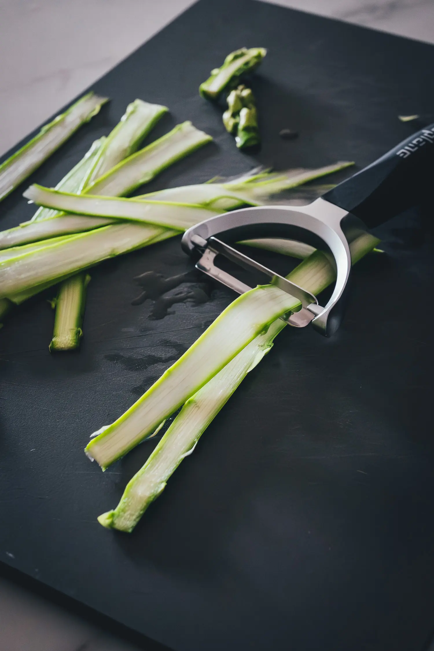 Roher grüner Spargel auf einem Schneidebrett, wird mit einem Sparschäler in feine Streifen gehobelt